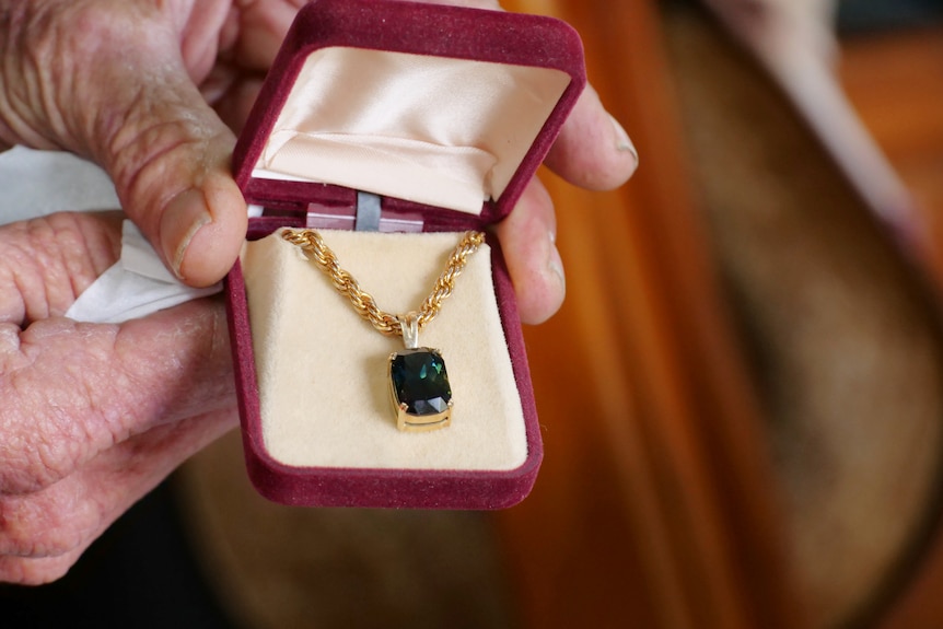 Murray's hands close up holding a jewellery box with a gold necklace and a big rectangle gem.