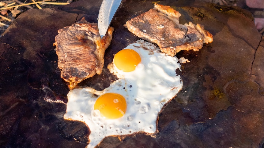Eggs and thick bacon are seen on a wooden slab being poked with a knife. A man bends down behind.