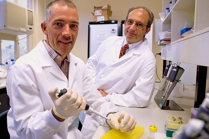 two men wearing white coats in a science lab. One is holding a test tube.