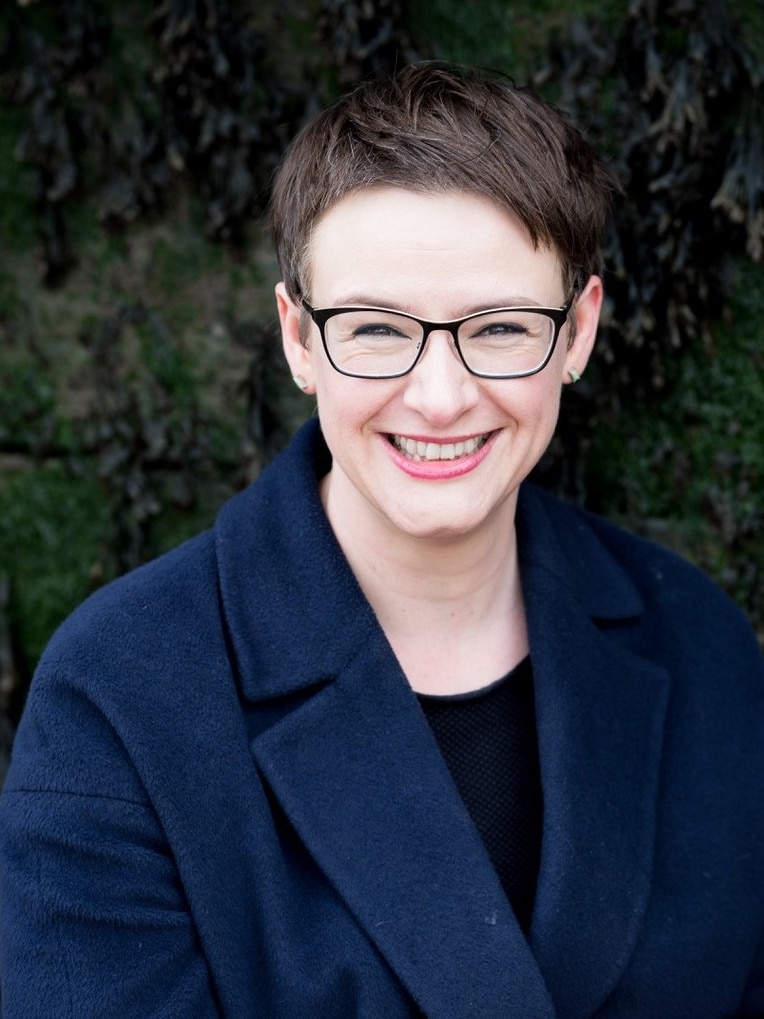 A portrait shot of a woman with a mousy brown pixie haircut and black rimmed glasses smiles