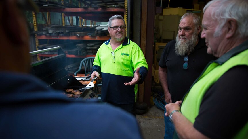 Four men chat as they stand around a barbecue, one with a meat flipper in his hand, planks of wood in the background.