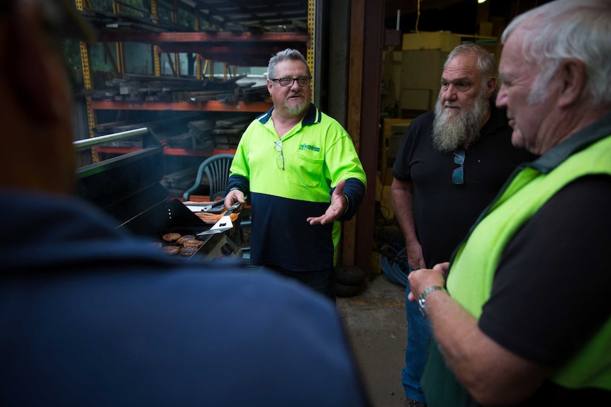 Four men chat as they stand around a barbecue, one with a meat flipper in his hand, planks of wood in the background.