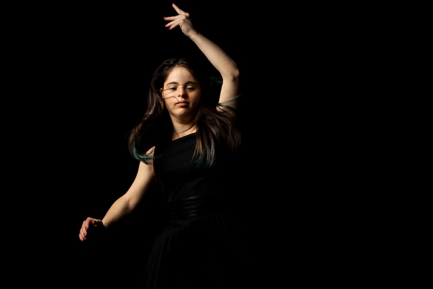 A white woman with long brown hair and blue tips, who also has down syndrome, dances fluidly in a darkened space.