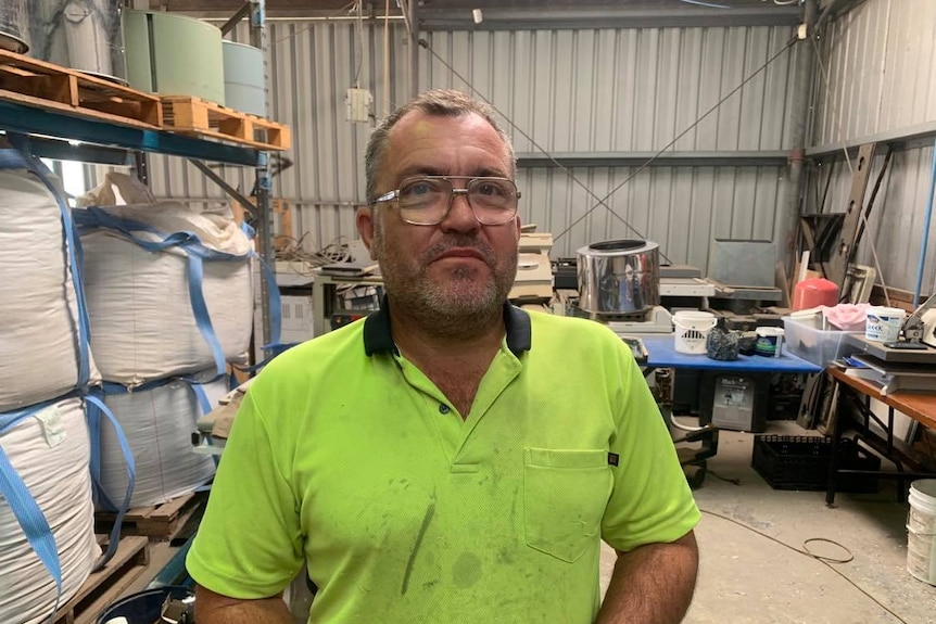 Michael Barker wears large clear glasses, a fluro yellow shirt and stands in a shed surrounded by pieces of recycling.