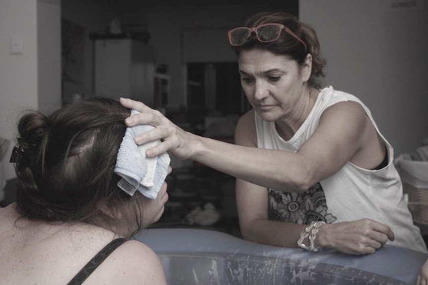 Renee Adair looking very still and focussed holds a wet flannel over the forehead of a woman in a birthing pool