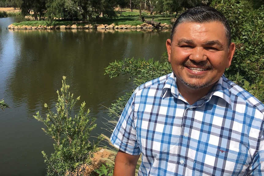 A man smiles as he stands on a riverbank in front of a river.