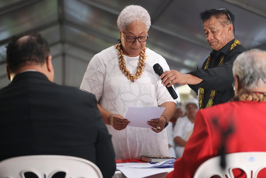 Woman in white outfit, glasses and necklace reads from paper next to man who holds up microphone.