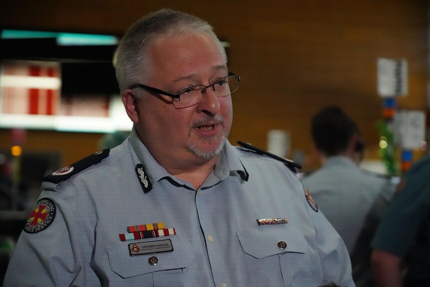 A middle-aged, bespectacled man with short grey hair and a neat beard, dressed in an ambulance officer's uniform.