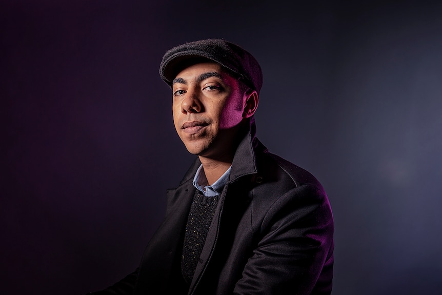 A man sits in front of grey backdrop with purple light on side of face in newsboy cap, shirt, jumper, coat with popped collar.