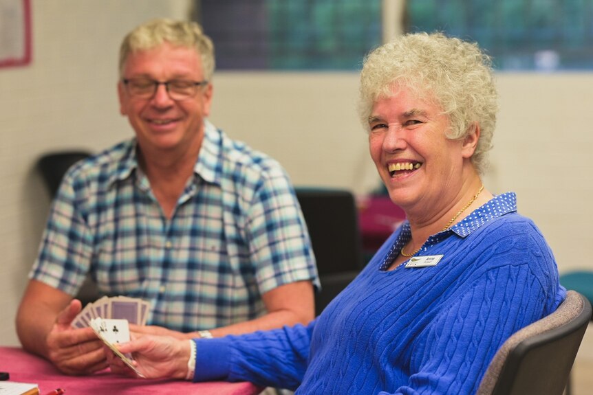 A woman with curly grey hair smiles into the camera while playing cards. 