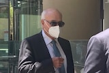 Two men in suits, ties and sunglasses walk outside a building, with one of them wearing a face mask.