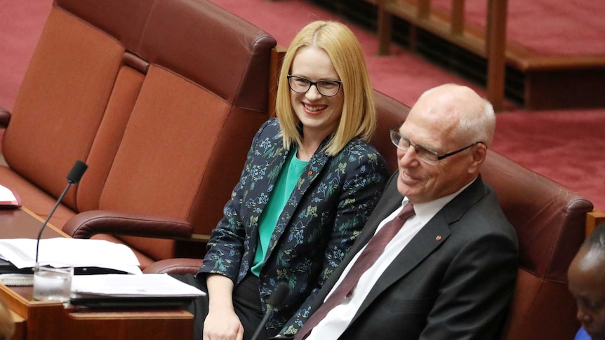 Stoker is smiling wearing dark rimmed glasses on the last row of the backbench.