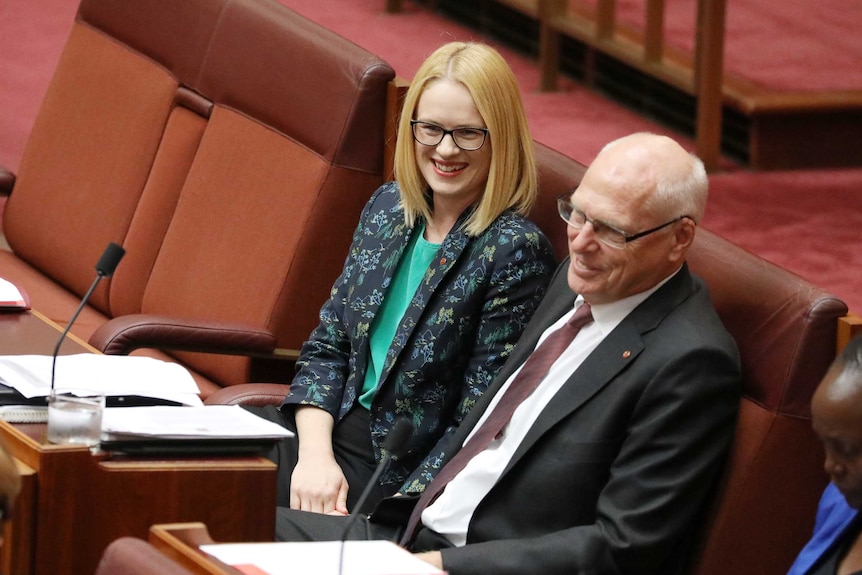 Stoker is smiling wearing dark rimmed glasses sitting next to Molan on the last row of the backbench.