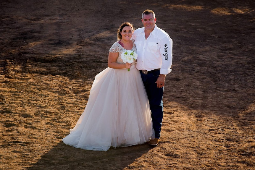 A woman wearing an elaborate white wedding dress stands on the dirt ground inside a rodeo ring with her husband.
