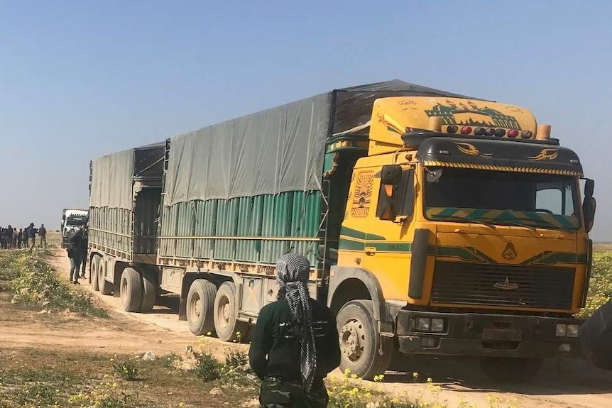 A large truck sits on the road, a military truck near it an a group of people stand nearby in a field.