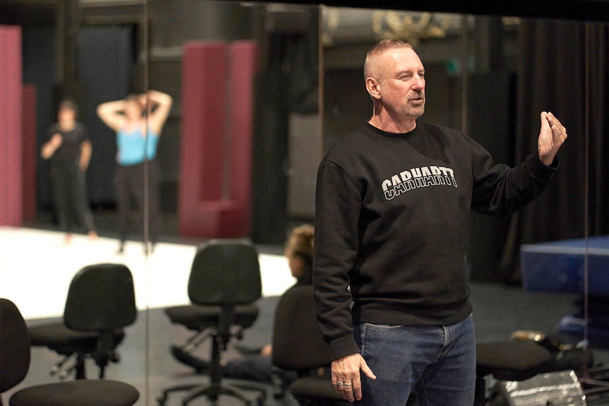 A man holds his hand out while mirrors in the background show rehearsing dancers