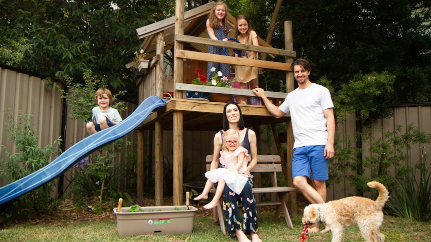 Four children, two parents and a dog sit on a wooden backyard playground.