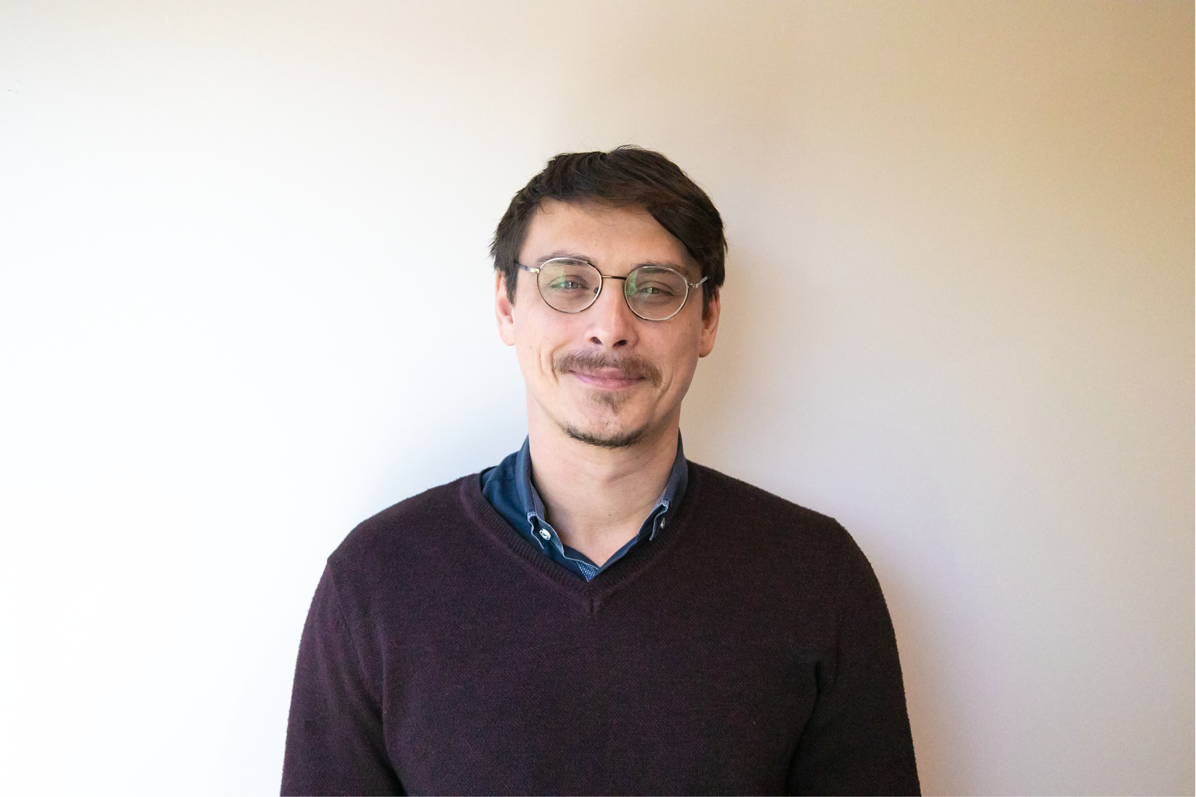 A man in a marron sweater and glasses standing in front of a white wall. 
