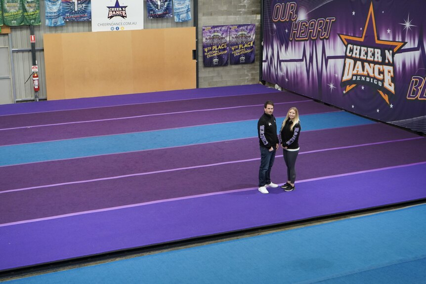 A man and woman, both dressed in company tracksuits, stand in deserted gymnasium usually filled by dancers and cheerleaders 