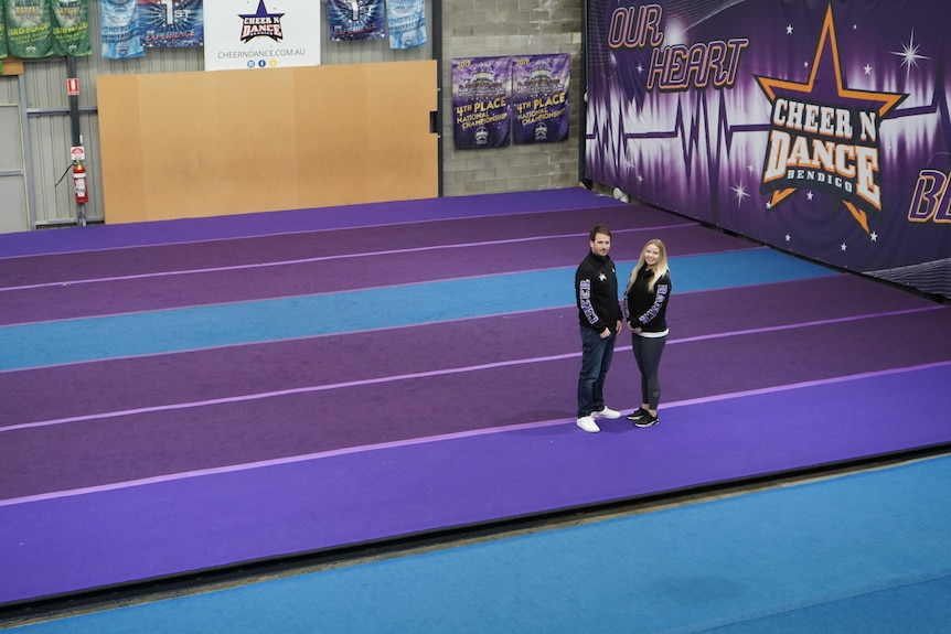 A man and woman, both dressed in company tracksuits, stand in deserted gymnasium usually filled by dancers and cheerleaders 