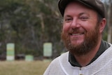 Man with Orange beard and flat cap stands in front of bee hives