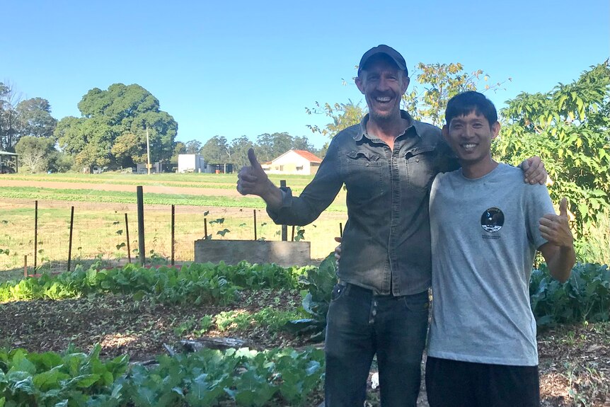 Two men stand next to each other with their thumbs up in a garden