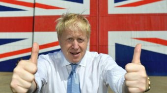 A man with blond hair gives a thumbs up to the camera