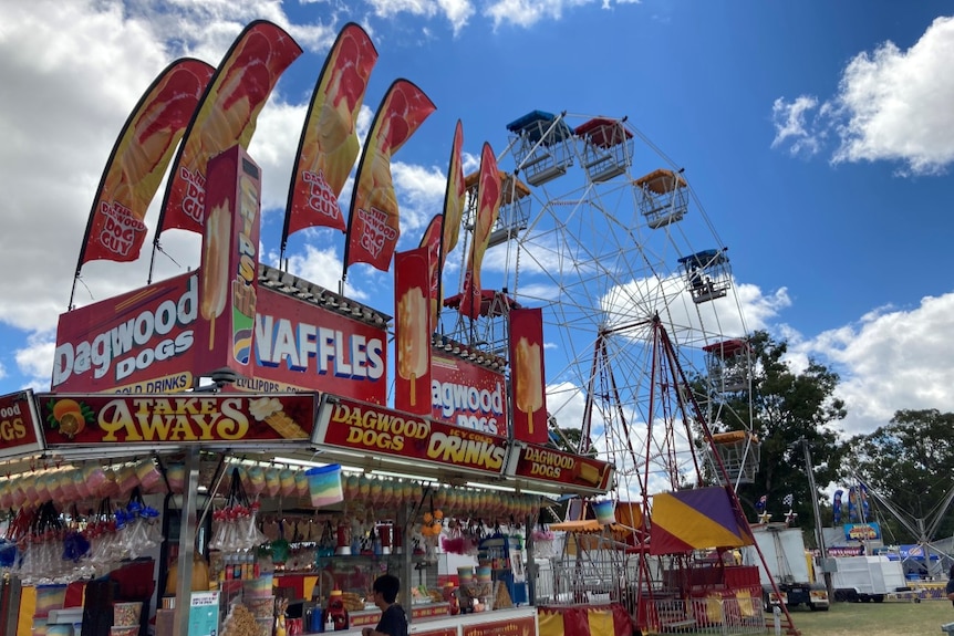 A dagwood dog stand in the foreground with a ferris wheel in the background at an agricultural show.