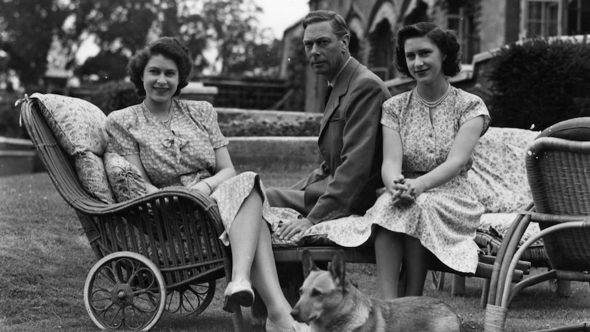 King George VI sitting on a chair with his daughters sitting either side of him.