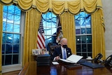 US President Joe Biden sits at the Resolute Desk with gold curtains behind him.