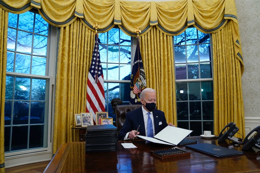 US President Joe Biden sits at the Resolute Desk with gold curtains behind him.