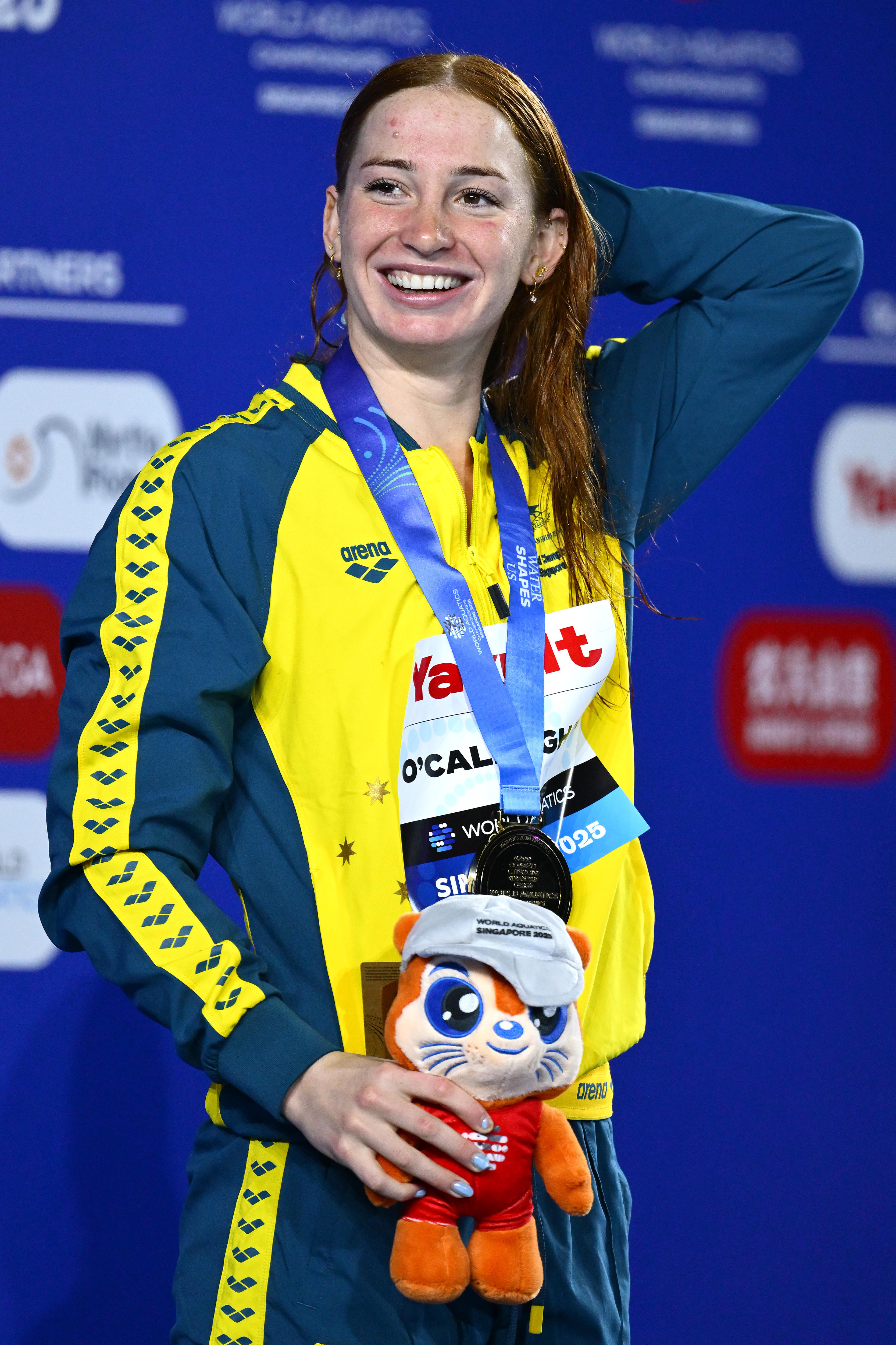 A smiling young woman with long, dark hair wears a brightly-coloured tracksuit and a medal.
