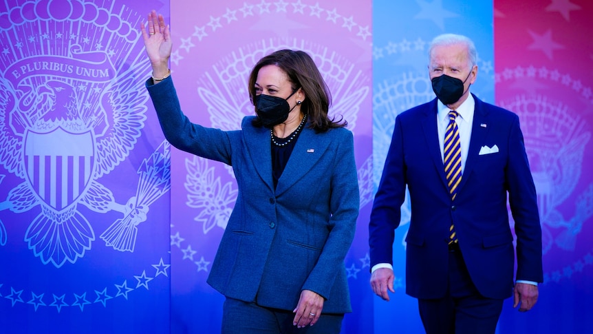 Kamala Harris in a navy pantsuit and black mask waves while Joe Biden walks behind her