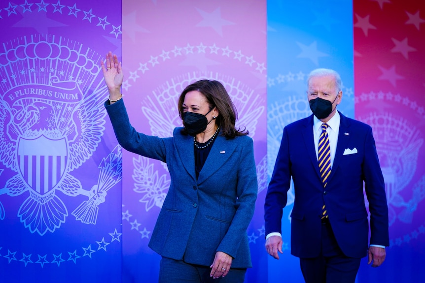 Kamala Harris in a navy pantsuit and black mask waves while Joe Biden walks behind her