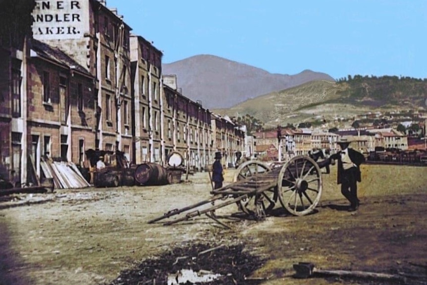 A man stands next to a cart, buildings to his right and a mountain in the distance