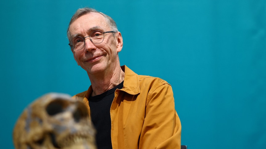 A middle-aged man wearing glasses smiles next to an old skull.