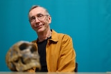 A middle-aged man wearing glasses smiles next to an old skull.