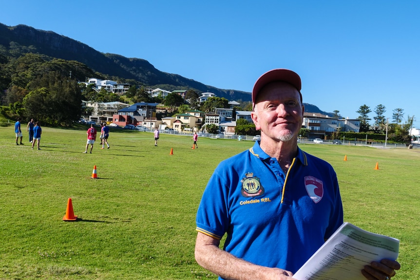 A man stands in front of a soccer field holding a grant application