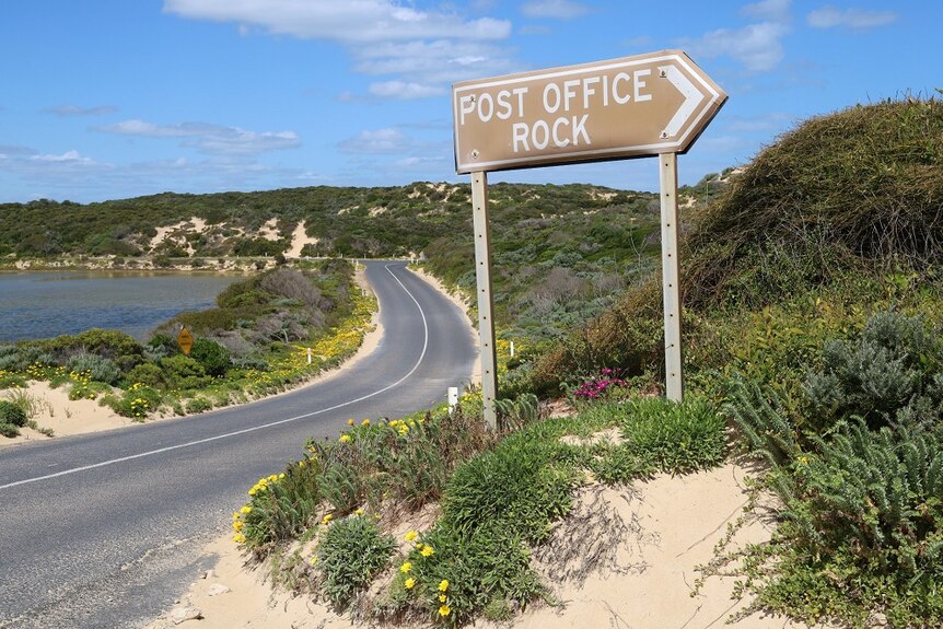 A sign reading 'Post Office Rock with a road and bay behind it.