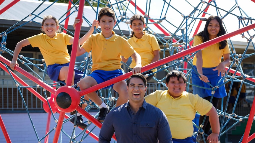 A man in a blue shirt poses for a photo with five primary school kids perched in a climbing apparatus