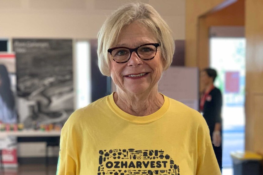 Older woman with grey hair wearing a yellow Oz Harvest shirt smiles while holding a brown sack of potatoes