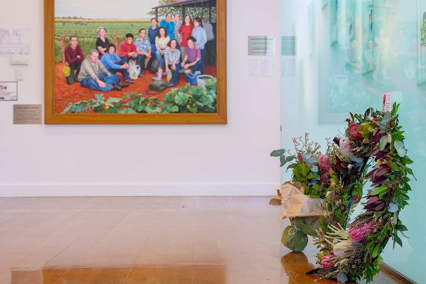 Several floral wreaths laid on the ground with a large painting of farm workers in the background.