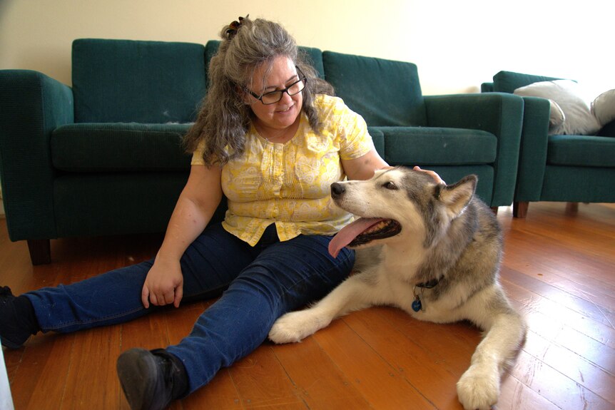 A woman in a yellow shirt sits on the floor of a living room with an elderly husky by her side