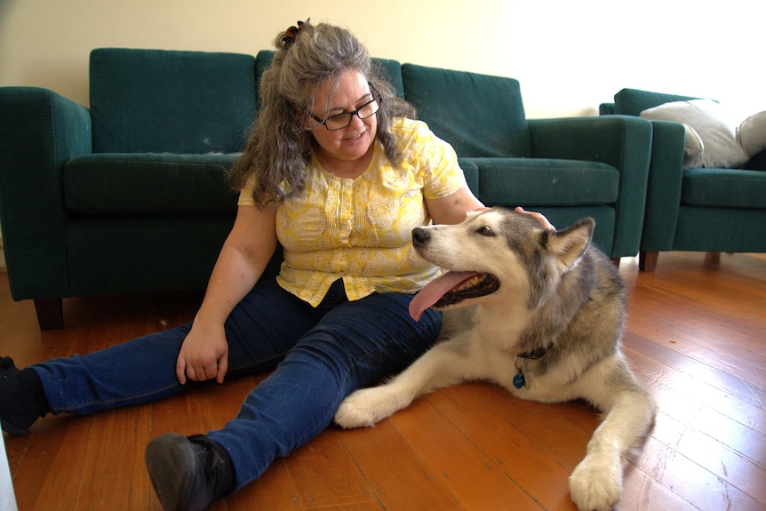 Una mujer con una camisa amarilla sentada en el suelo de una sala de estar con un anciano husky a su lado