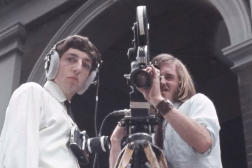 A man wearing headphones looks at the camera, while another men looks through a video camera lens.
