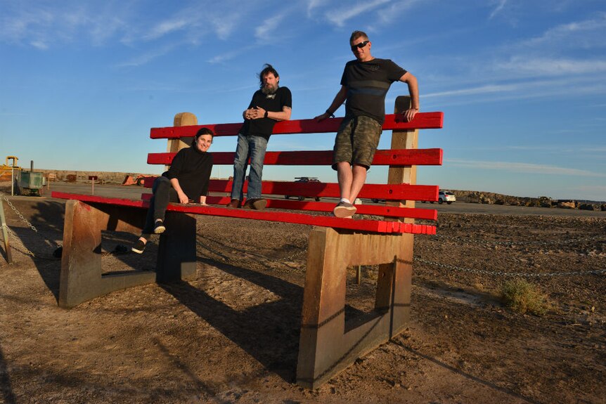 Prue Adams sitting, Tony Hill and Carl Saville standing on giant red seat on sparse landscape.
