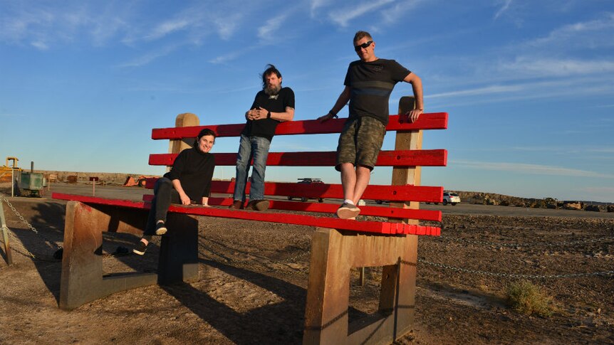 Prue Adams sitting, Tony Hill and Carl Saville standing on giant red seat on sparse landscape.