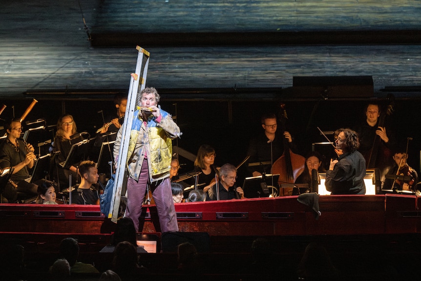 A white middle-aged man performs on stage in front of an orchestra, wearing painter's clothes and carrying a ladder & harmonica.