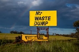 A tractor with a sign that reads 'No Waste Dump' is parked in a wheat field. A rainbow is in the sky.