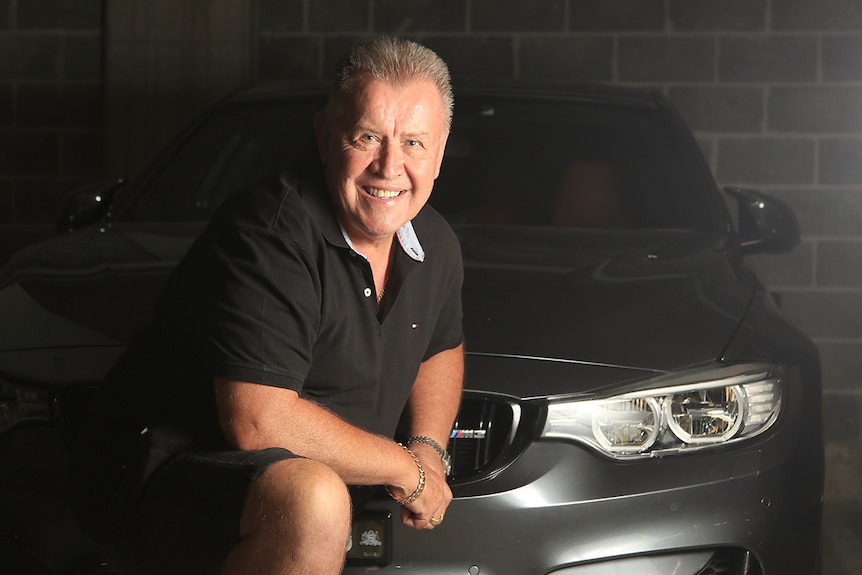 Ian Luff crouches down next to the front of a grey sports car in a car park.